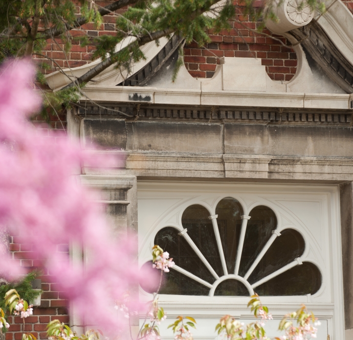 Pink blossoms and pine branches partially obscure a white door with a fanned glass design in an ornate stone doorway of a red brick building.
