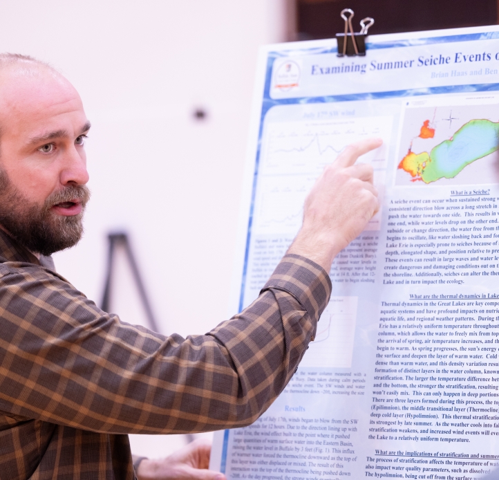 A bearded man presents a research poster, pointing to a colorful map of Lake Erie while explaining data to an audience member. The poster discusses summer seiche events on Lake Erie, with charts and diagrams visible.