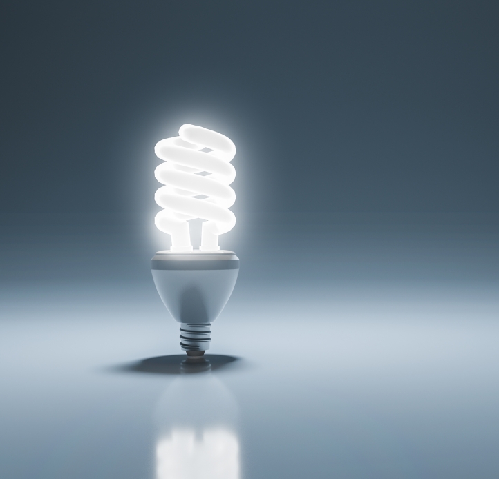  A single glowing spiral compact fluorescent light (CFL) bulb stands on a reflective surface against a dark background, with ample copy space to the right.