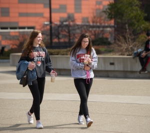  Two female college students walk across a campus paved area on a sunny day, both wearing leggings and Buffalo State sweaters, holding drinks.