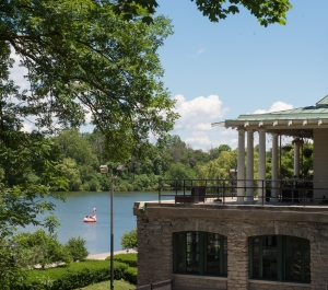 Stone building with green roof and pillars overlooks a pond with a pink flamingo float, surrounded by trees under a partly cloudy sky.