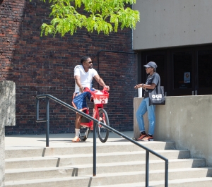  A man on a red bicycle with a basket talks to another person standing on a set of concrete stairs in front of a brick building.