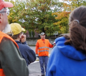 A man wearing a bright orange safety vest speaks to a small group of people gathered outdoors, during a campus or community event. Several listeners in the foreground wear jackets and hats while standing in a parking lot area. Behind the speaker are trees showing fall colors of green, yellow, and orange, suggesting an autumn setting.